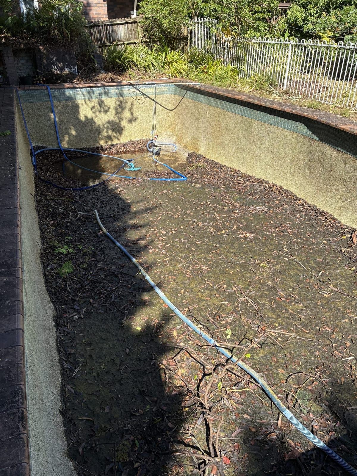 Drained pebblecrete pool with leaf litter across the floor, drain hoses running out, part way through a clean-out