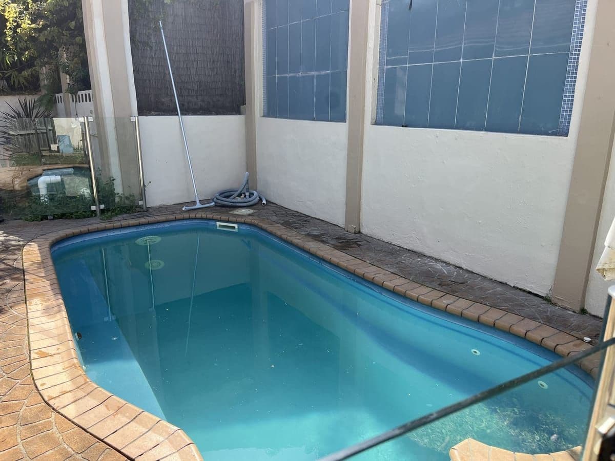 Narrow courtyard pool part way through a service, with brick paver coping, glass fencing and the vacuum hose on the edge