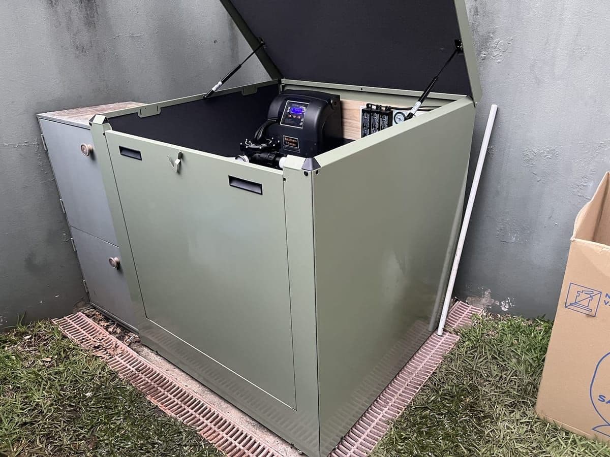 Olive-green acoustic equipment enclosure on a paver base, with the lid propped open to show the chlorinator inside