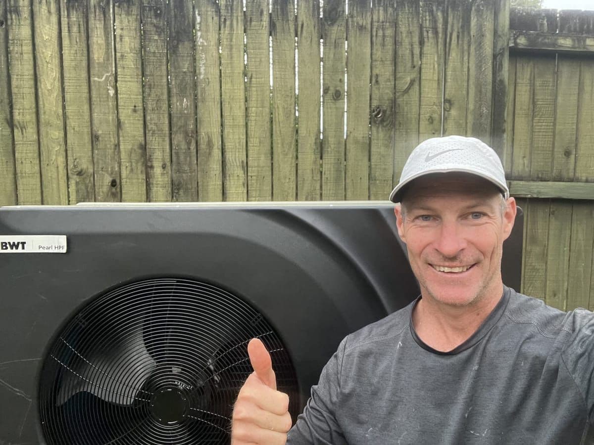 Andy standing beside a newly installed heat pump against a timber paling fence, giving a thumbs up